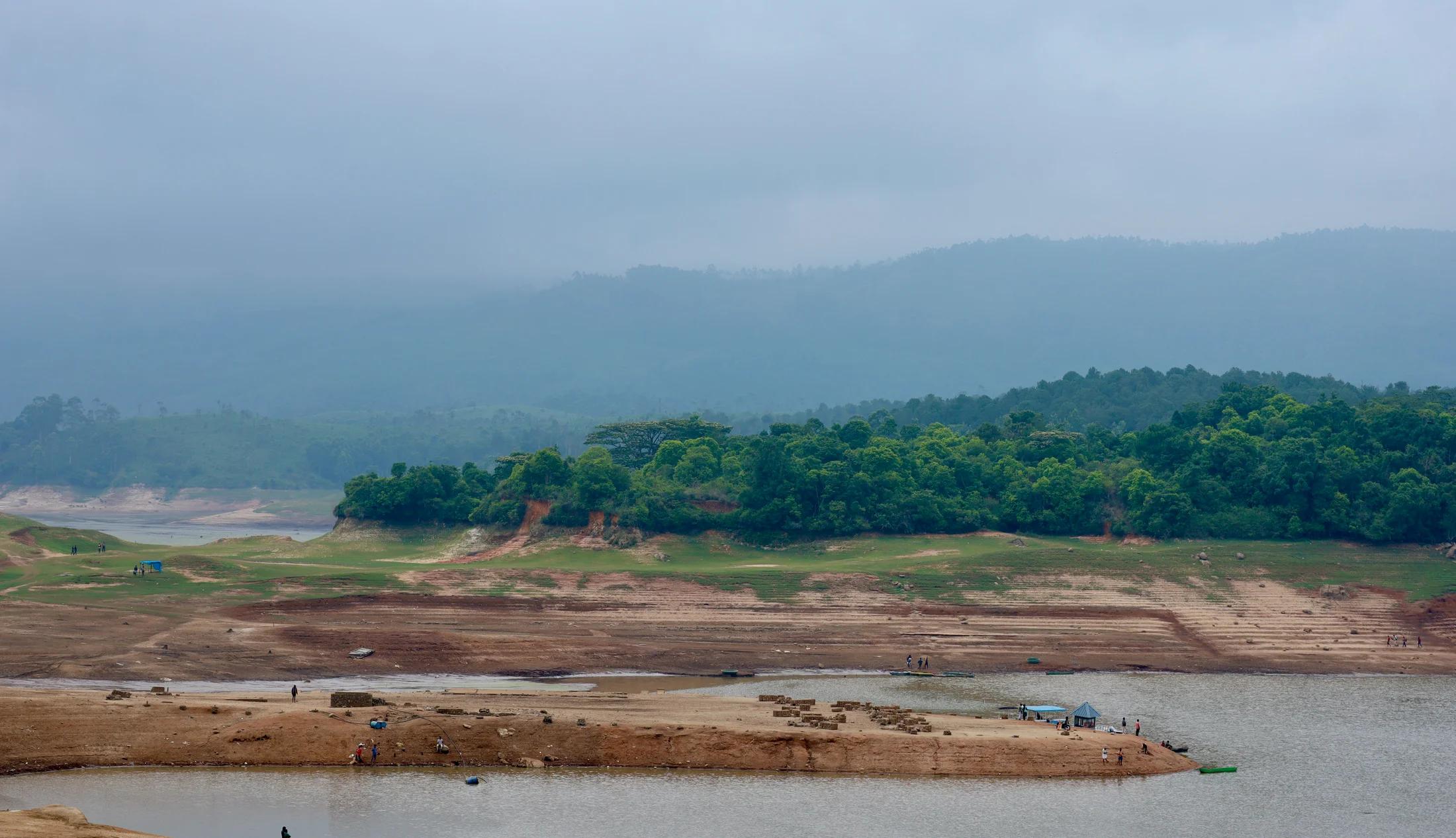A reservoir shoreline backed by green forest and misty hills in Kerala.