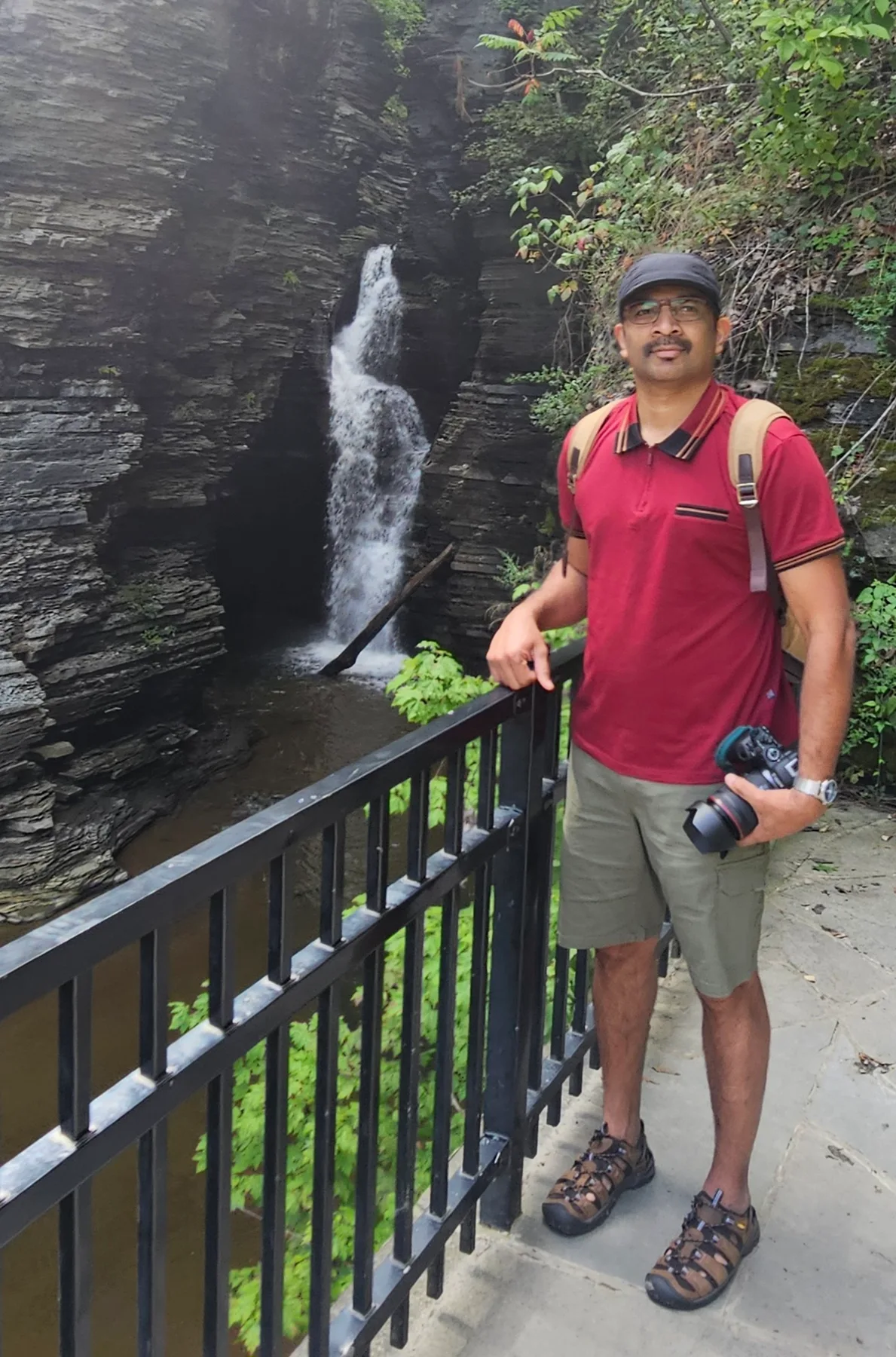 A man holding a camera beside a waterfall and stone gorge at Watkins Glen.